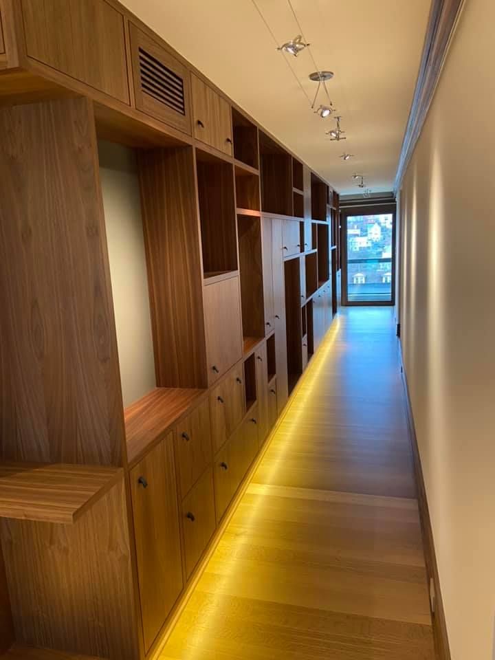 Wooden hallway with built-in lockers and cabinets, lit by recessed ceiling lights, leading to a bright windowed end.