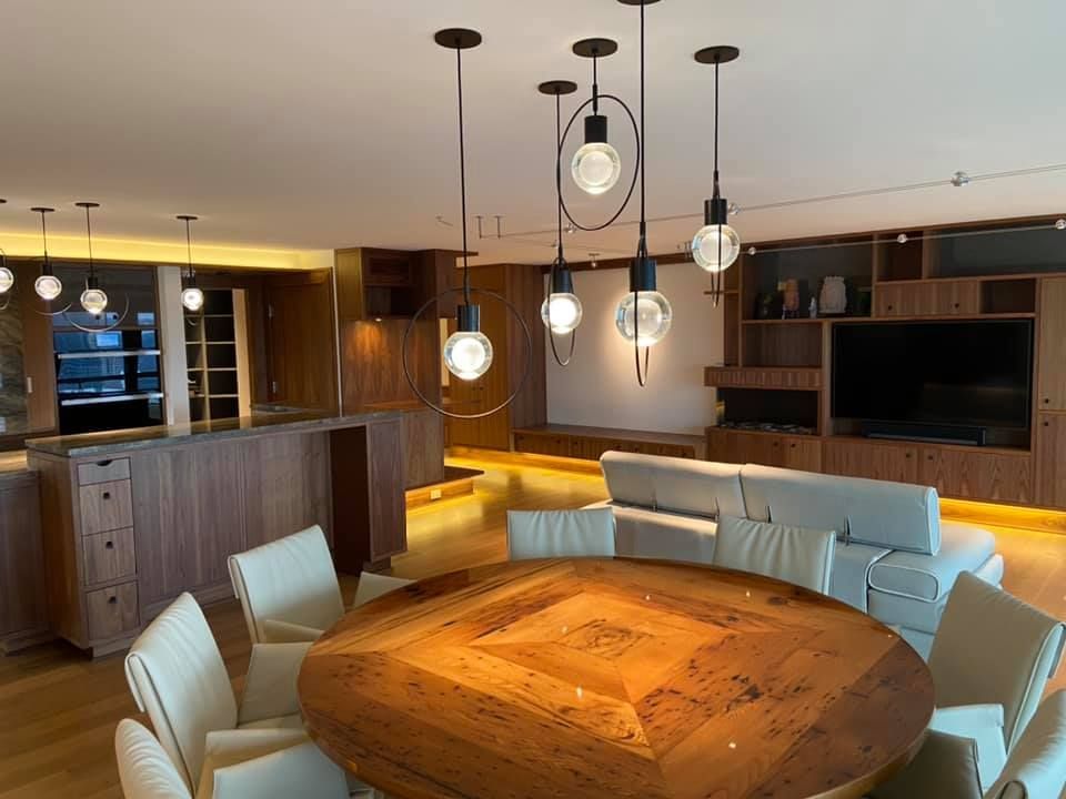 Modern dining room with a round wooden table, light chairs, pendant lights, and open kitchen bar.