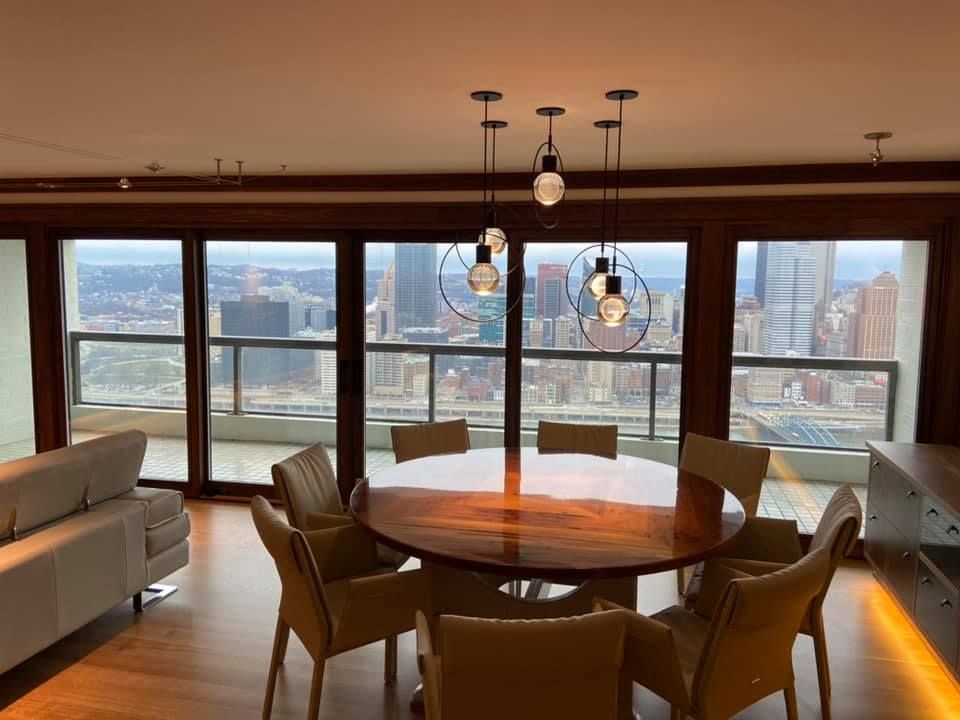 Dining room with round wooden table, chairs, sofa, and city view through large windows