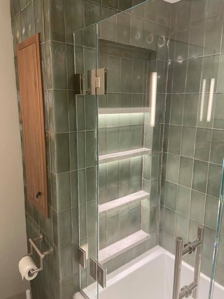 Green-tiled shower with built-in shelves, glass door, and wood accent wall in a bathroom