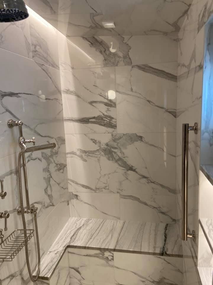 Modern marble-tiled shower with glass door, handheld showerhead, built-in bench, and wall niche.