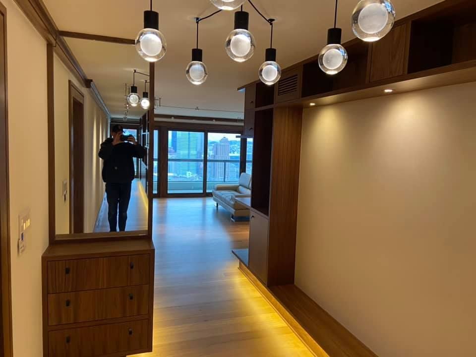 Modern hallway with hanging globe lights, wooden floor, beige walls, and large windows at the far end