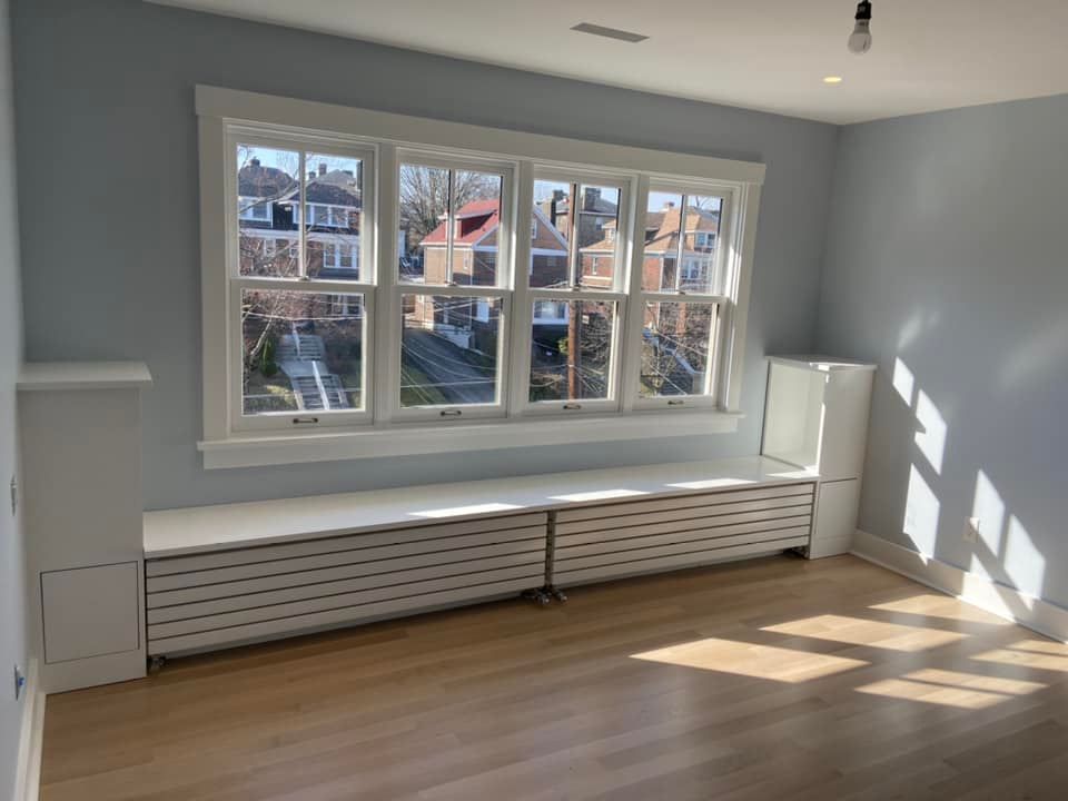 Empty sunlit room with gray walls, large windows, white radiator cover, and hardwood floor.