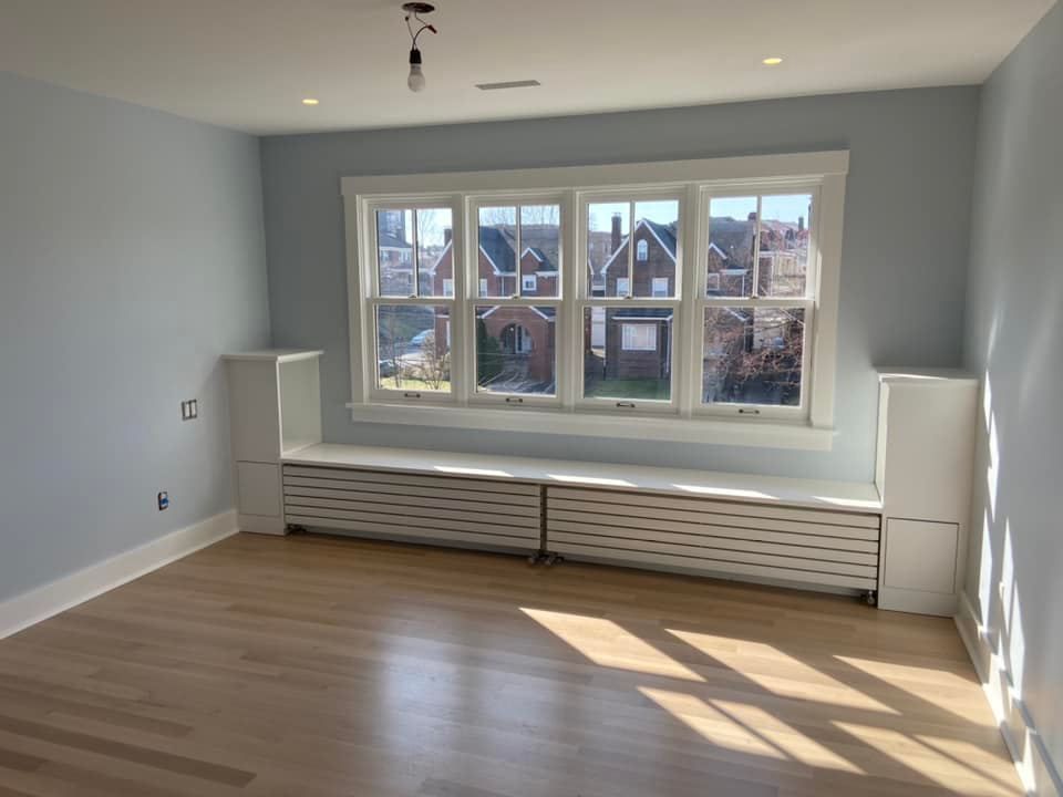 Empty living room with light wood floors, gray walls, and a large window above a radiator cover.