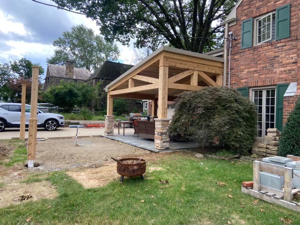 Covered patio under construction beside a brick house with wooden beams, stone posts, and a fire pit in the yard