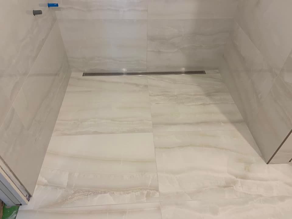 Empty tiled shower stall with beige marble-look walls and floor, drain at back wall.
