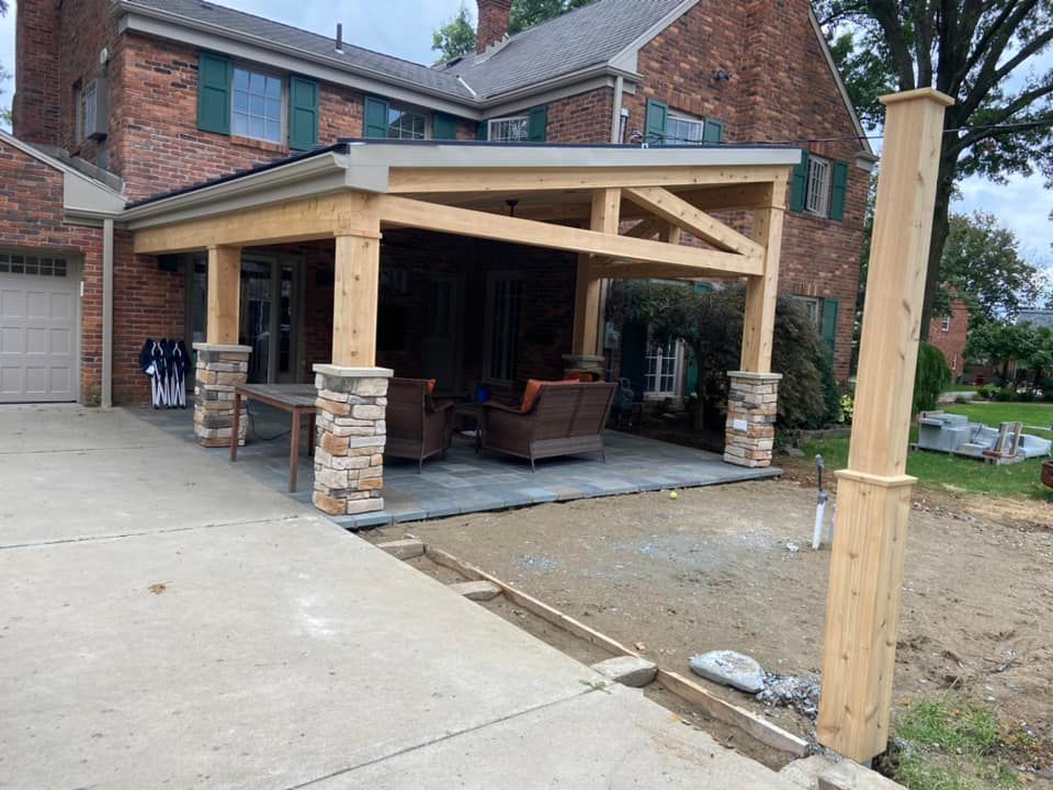 Partially built wooden patio roof with stone columns attached to a brick house and concrete driveway