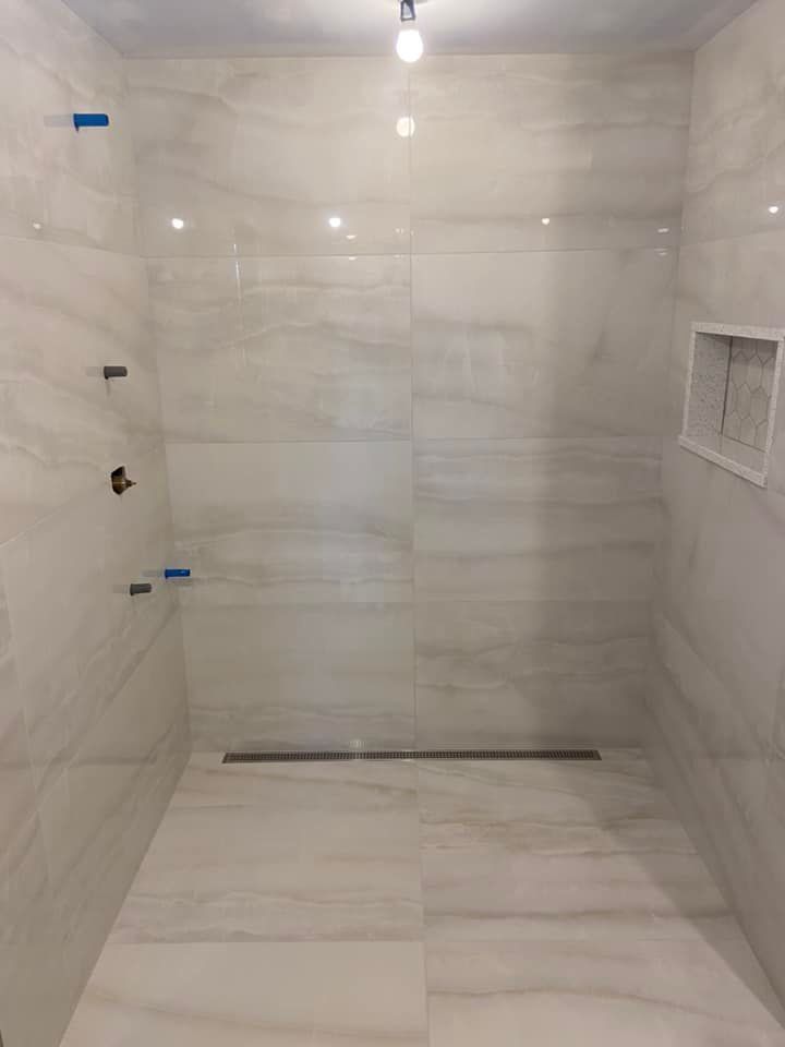 Empty tiled shower with white marble-look walls and floor, recessed shelf, and bare ceiling light.