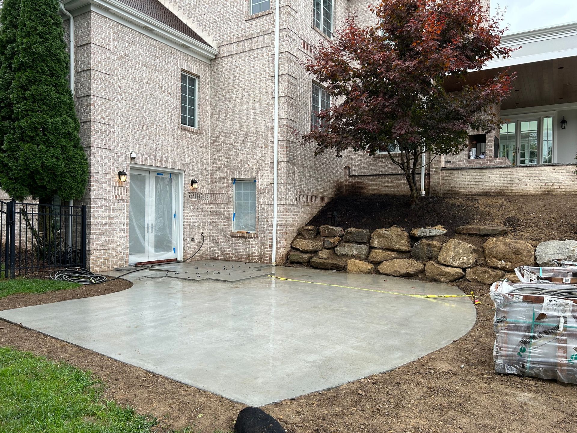 Back patio with fresh concrete slab beside a brick house and stone retaining wall.