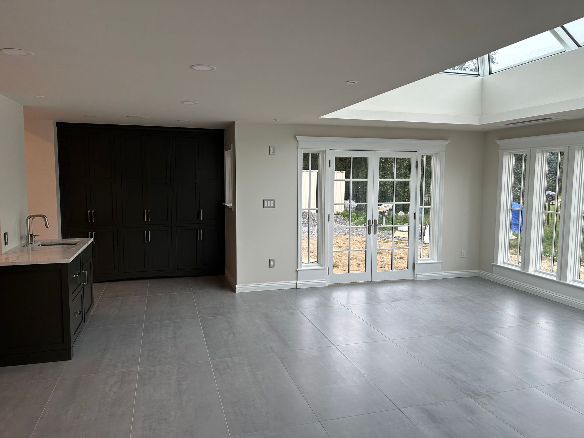 Empty modern kitchen and living area with gray floors, dark cabinets, and large glass doors to a patio