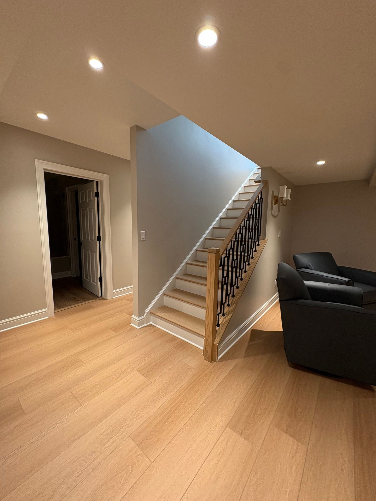 Empty modern living room with wood floors, stairs, recessed lights, and a dark sofa.