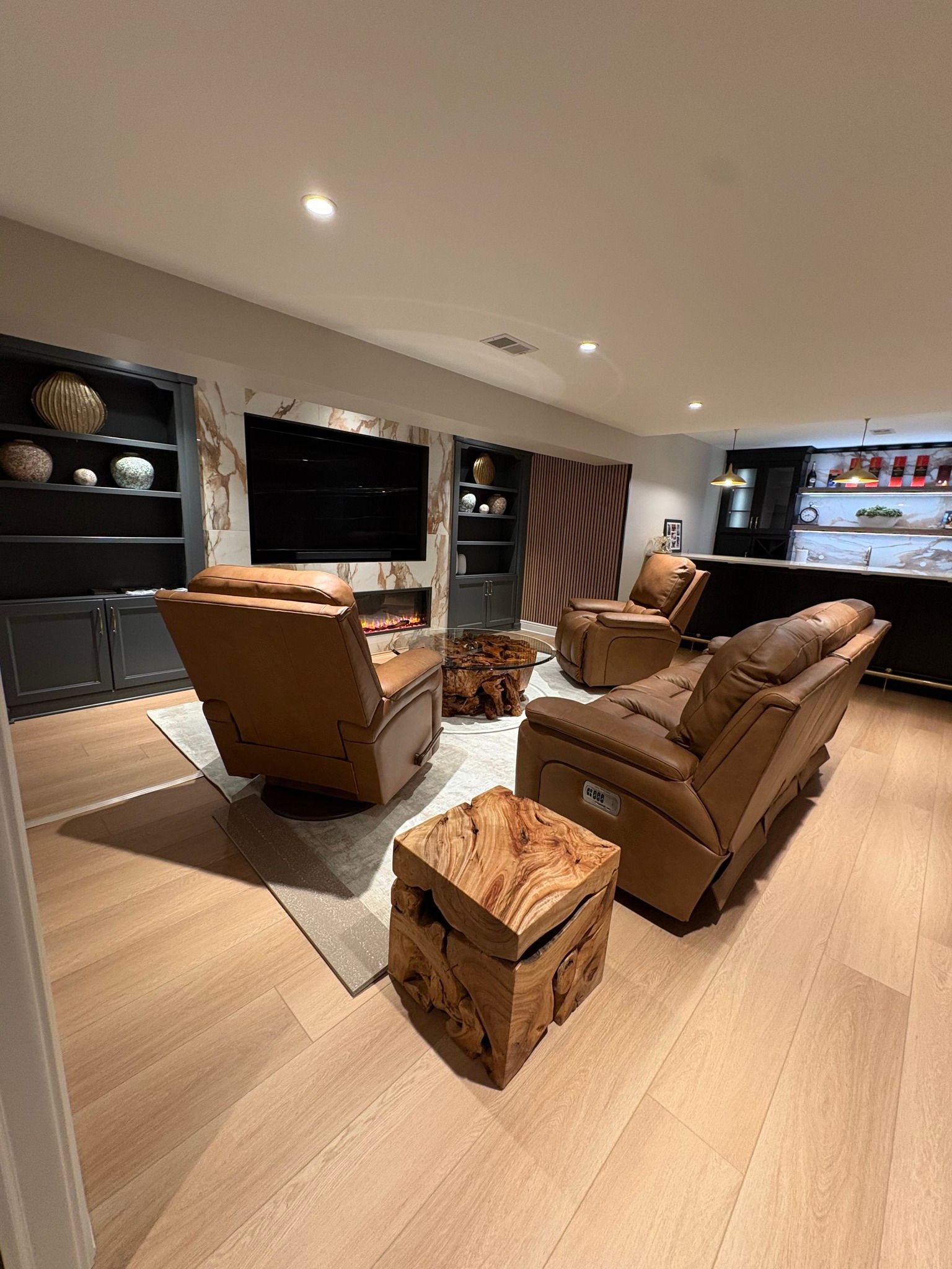 Modern living room with tan recliners, wall TV, fireplace, and built-in shelving under recessed lights