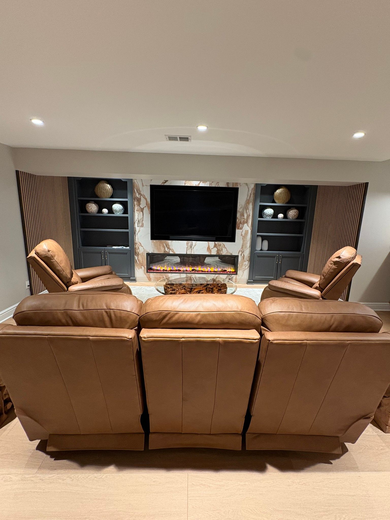Basement media room with tan recliners, built-in shelves, and a wall-mounted TV by a fireplace.