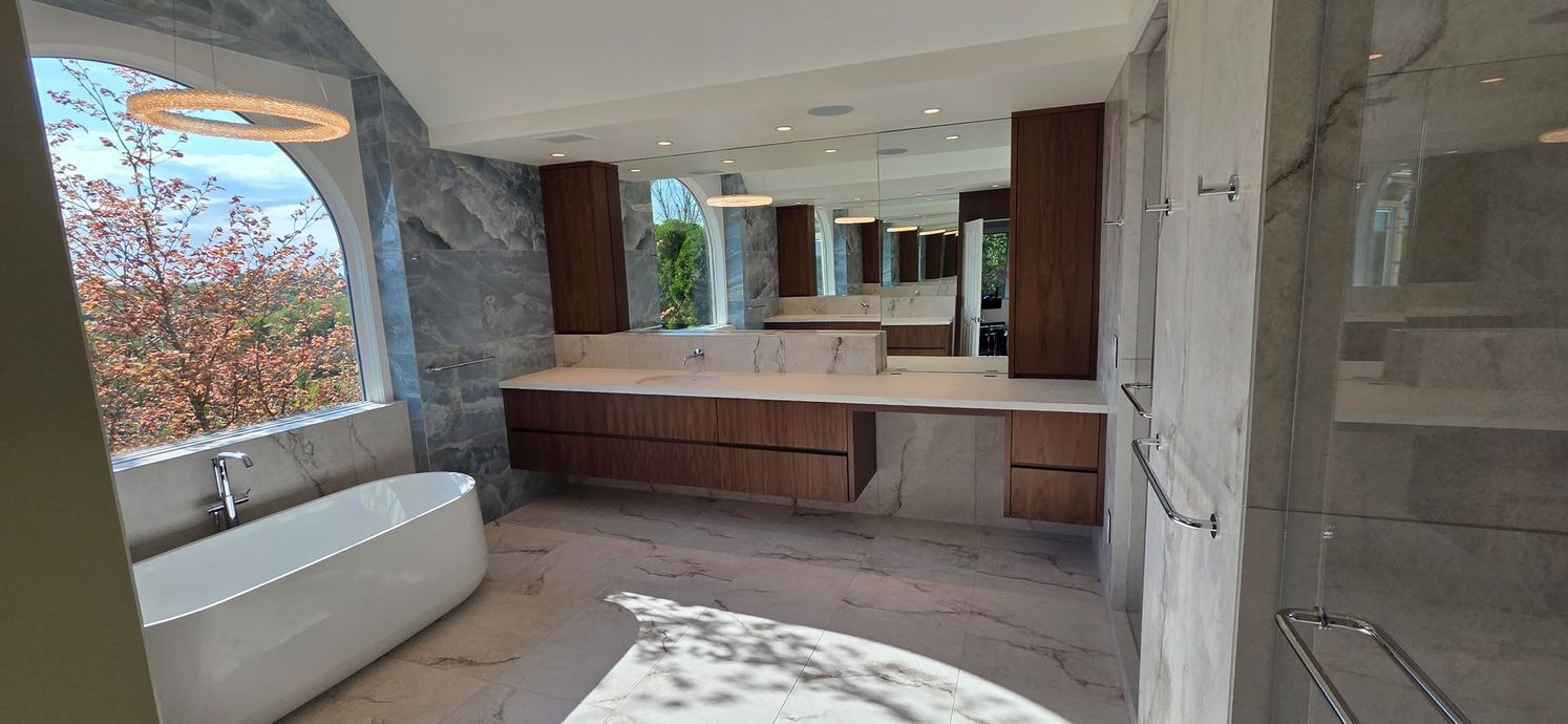 Modern bathroom with wooden vanity, large mirror, stone walls, and a freestanding tub near a window.