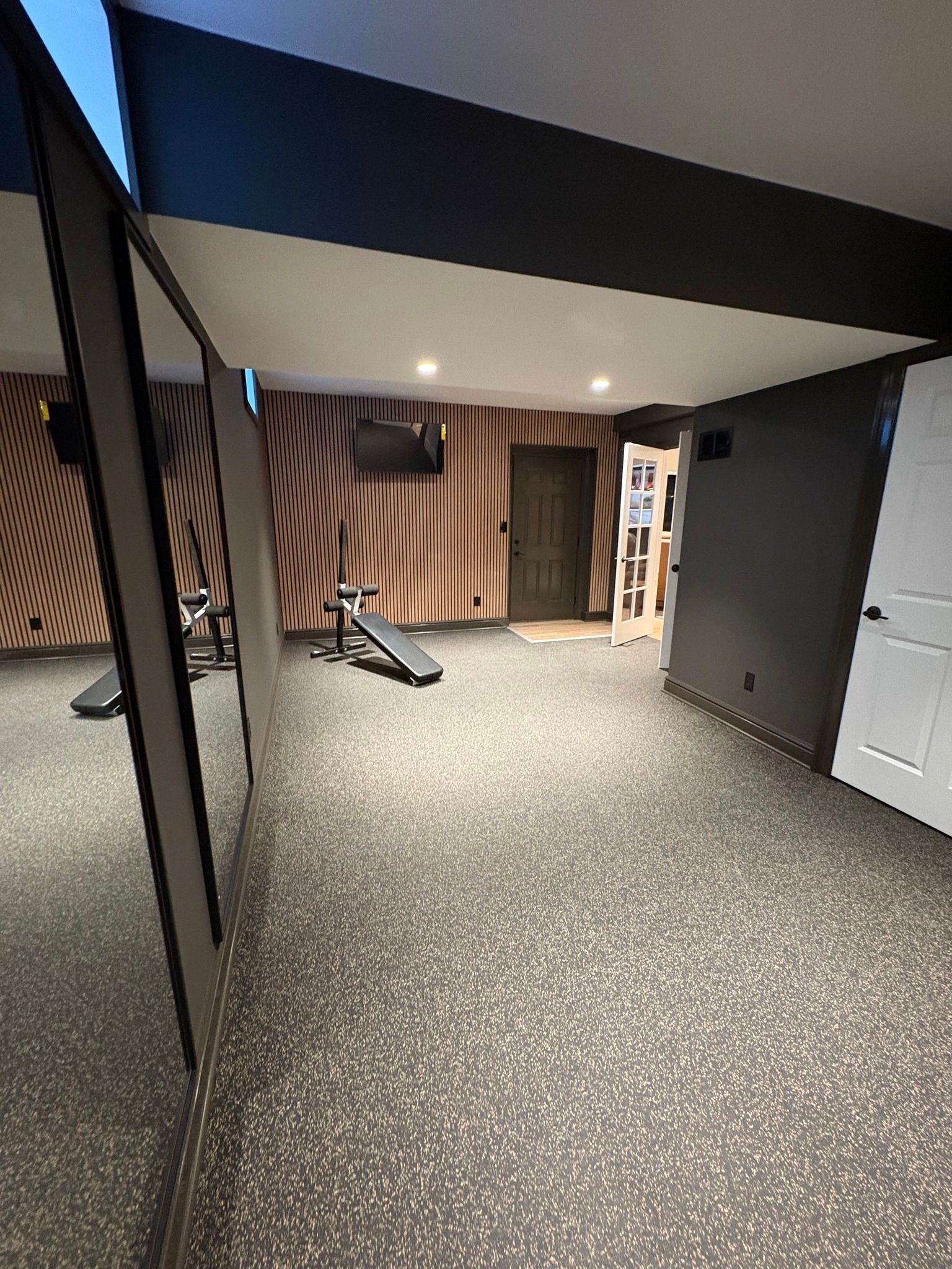 Empty basement room with speckled floor, dark ceiling, wood-paneled wall, and exercise equipment near the back.