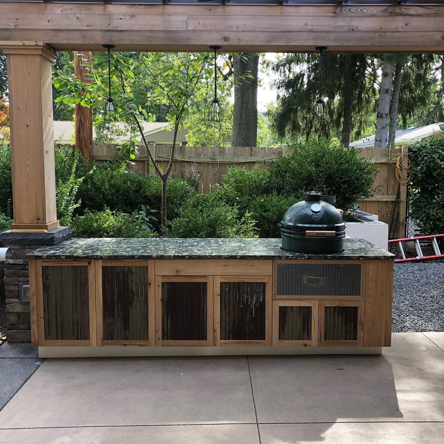 Outdoor stone counter with a green ceramic grill beneath a wooden pergola in a backyard patio