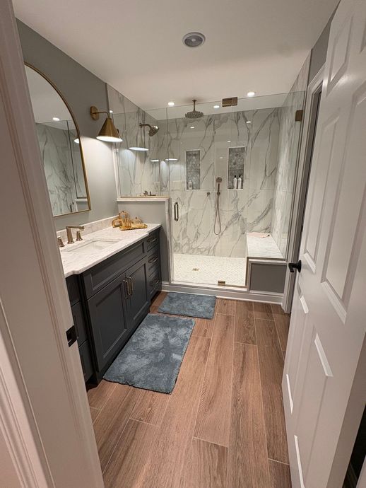 Narrow bathroom with marble shower, dark vanity, wood floor, and a white door open in the foreground