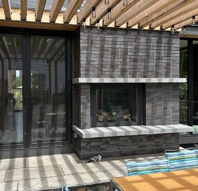 Outdoor stone fireplace beneath a slatted patio roof beside sliding glass doors and stacked materials on a deck.