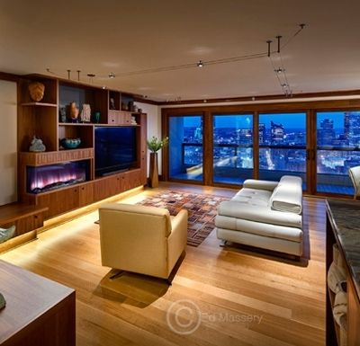 Modern living room with wood floors, white sofa, tan chair, built-in TV, and large city-view windows at dusk