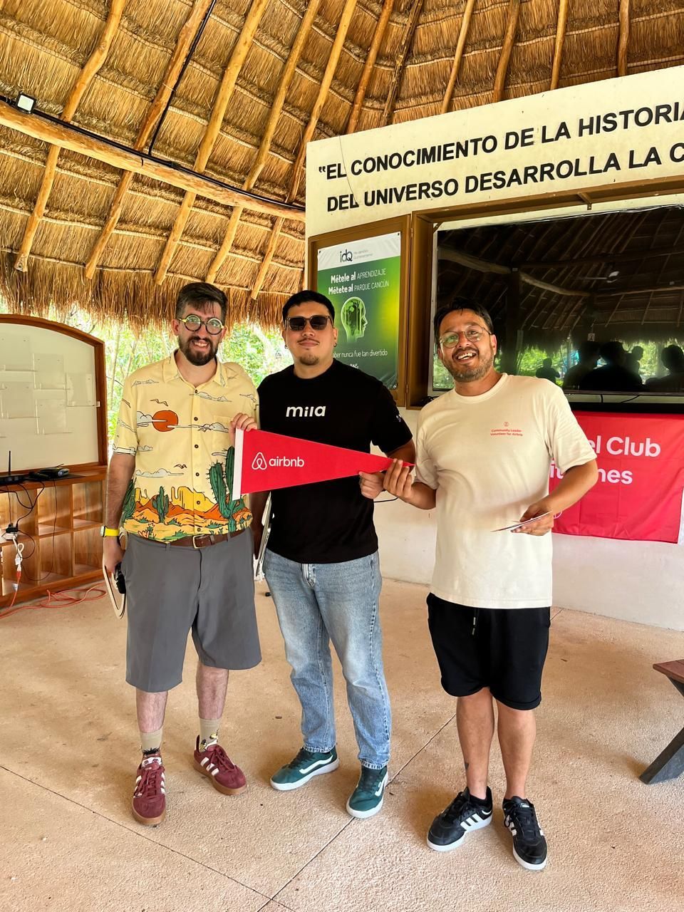 Tres personas posando en el interior de una casa con techo de paja, sosteniendo un cartel rojo con una bandera española detrás.