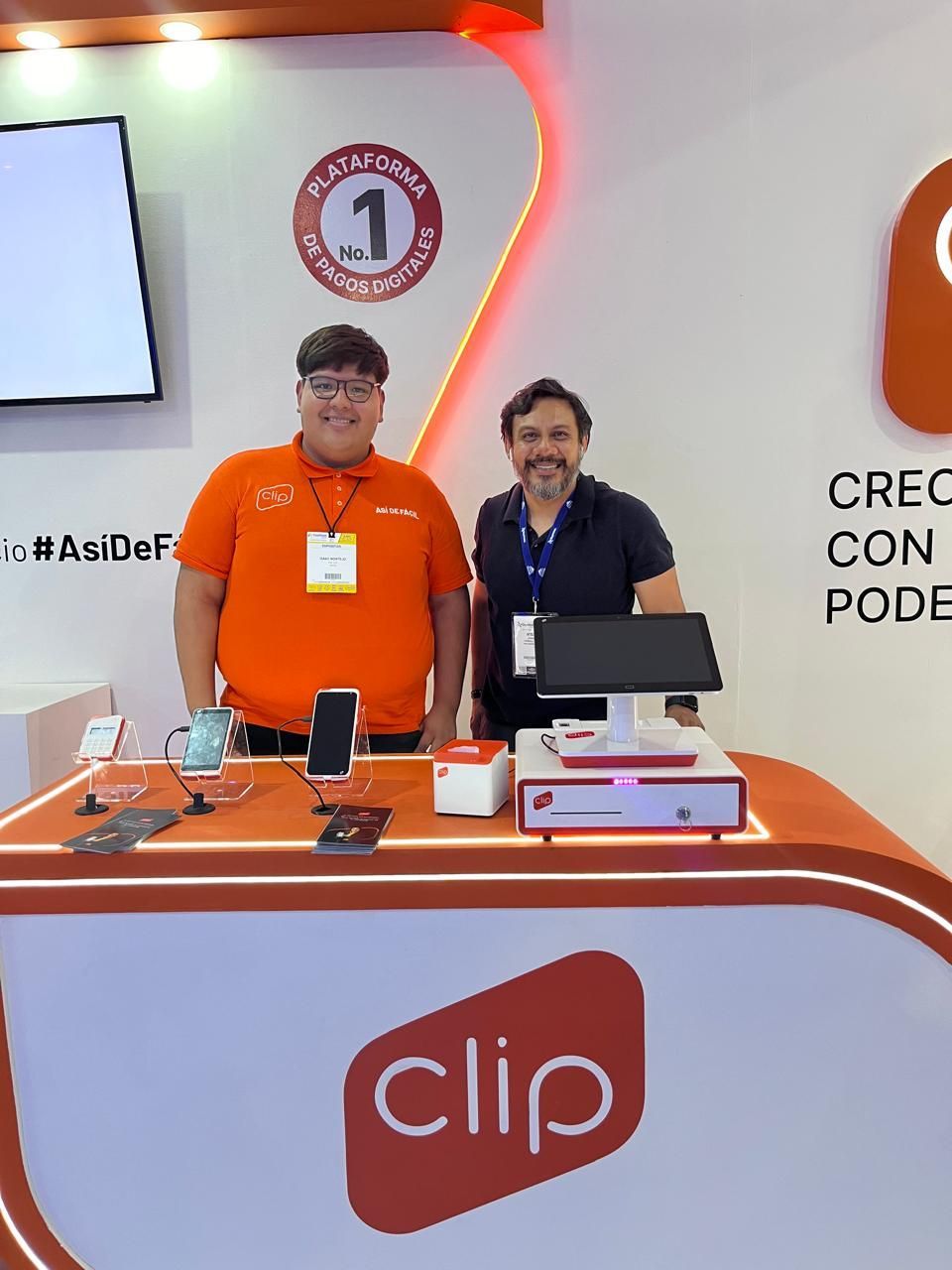 Dos personas detrás de un stand de Clio en una feria comercial, con ordenadores portátiles y la marca en color naranja.