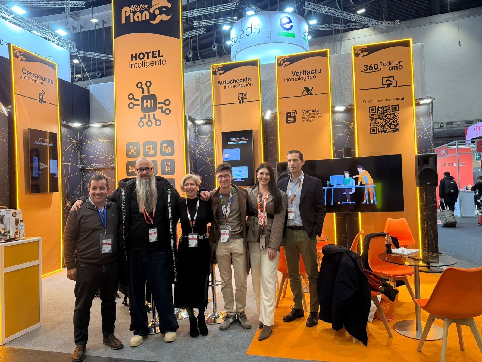 Grupo de personas posando en un stand de feria comercial de color naranja y azul con pancartas altas y sillas.