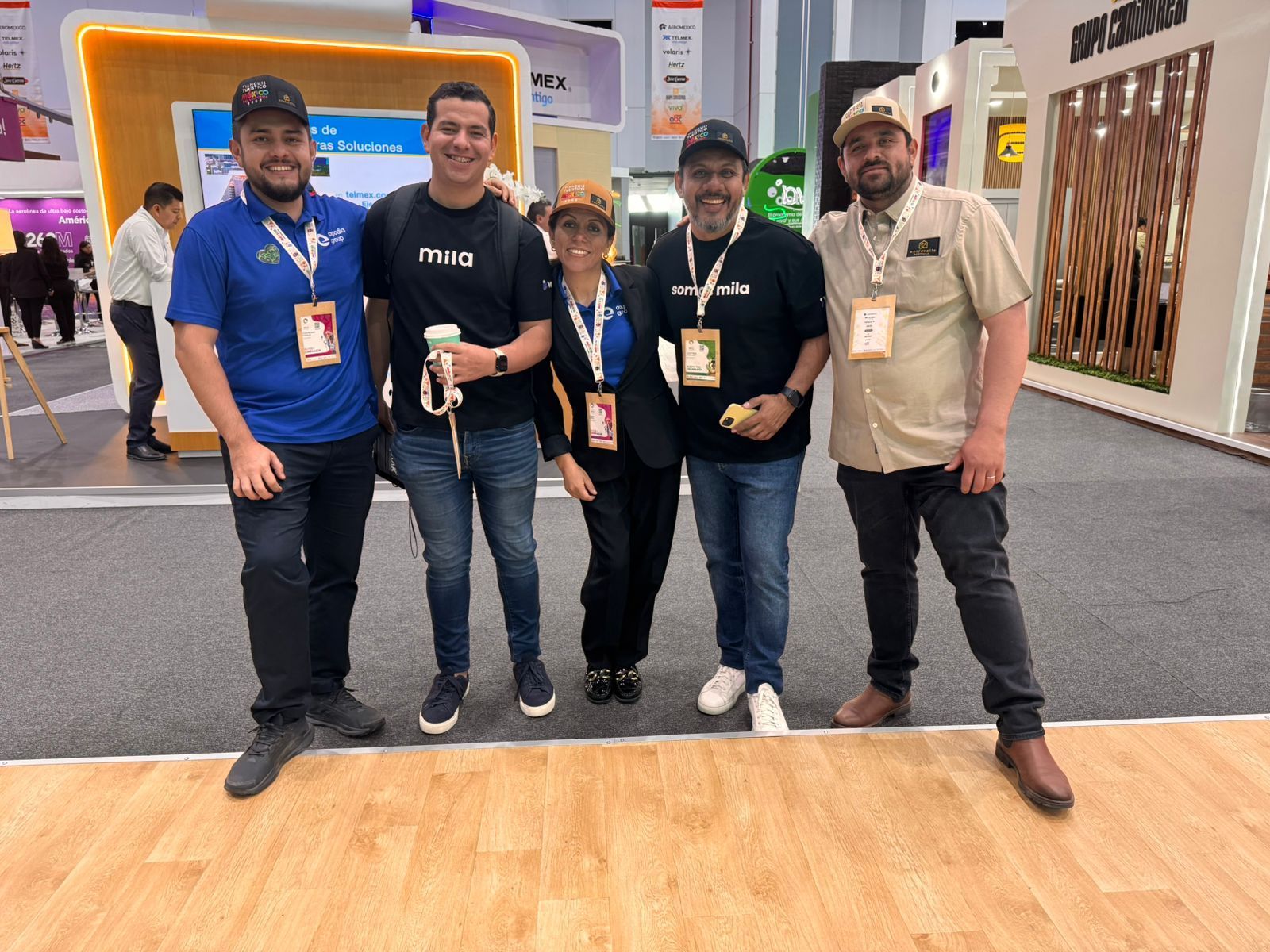 Cuatro personas posando en un stand de una feria tecnológica, sosteniendo bebidas y luciendo credenciales del evento.