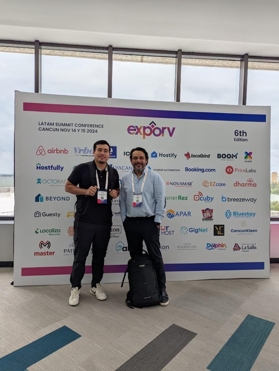 Dos personas posando frente a un telón de fondo de la exposición, cada una luciendo una acreditación del evento.