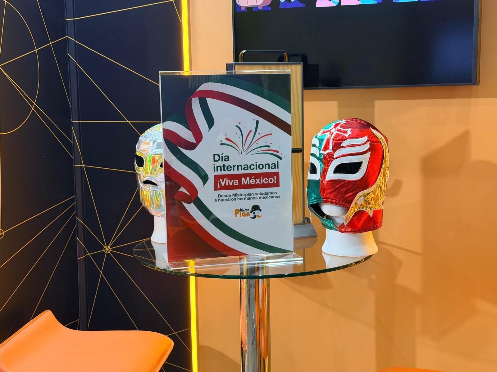 Tres coloridas máscaras de lucha libre expuestas sobre una mesa de cristal junto a una placa en un stand de exhibición.