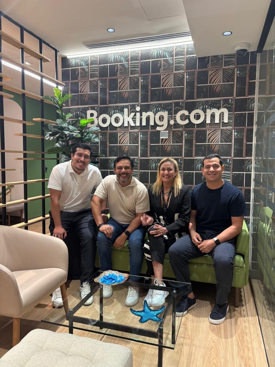 Cuatro personas sentadas en un vestíbulo, sonriendo junto a un cartel de Booking.com y una mesa de centro de cristal.