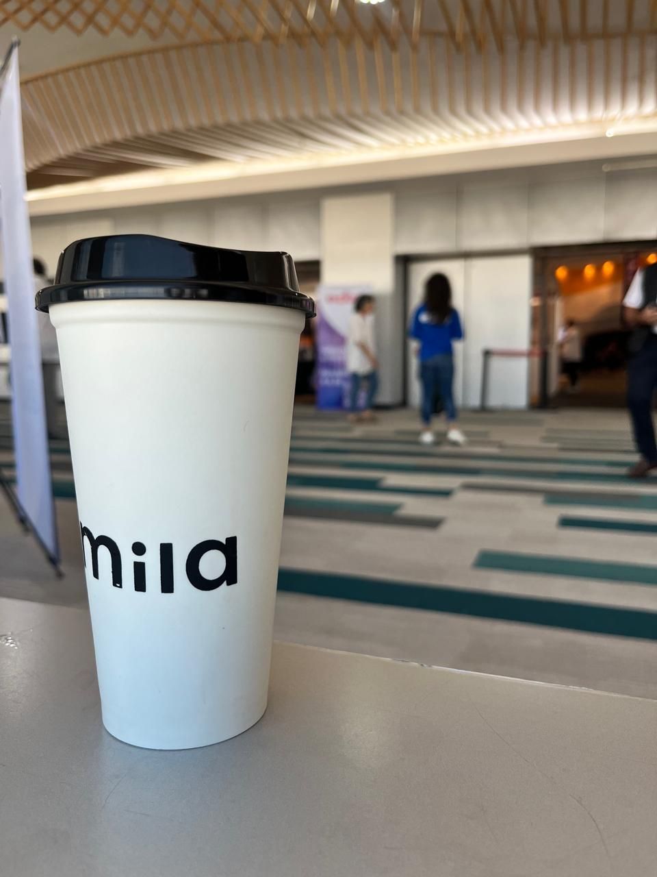 Vaso de café blanco para llevar con tapa negra sobre una mesa en un vestíbulo interior borroso.
