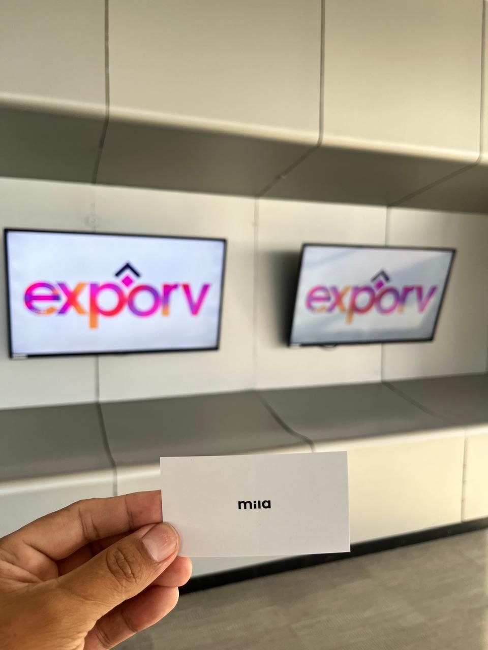 Una mano sostiene una tarjeta de presentación blanca frente a dos pantallas montadas en la pared que muestran el logotipo de