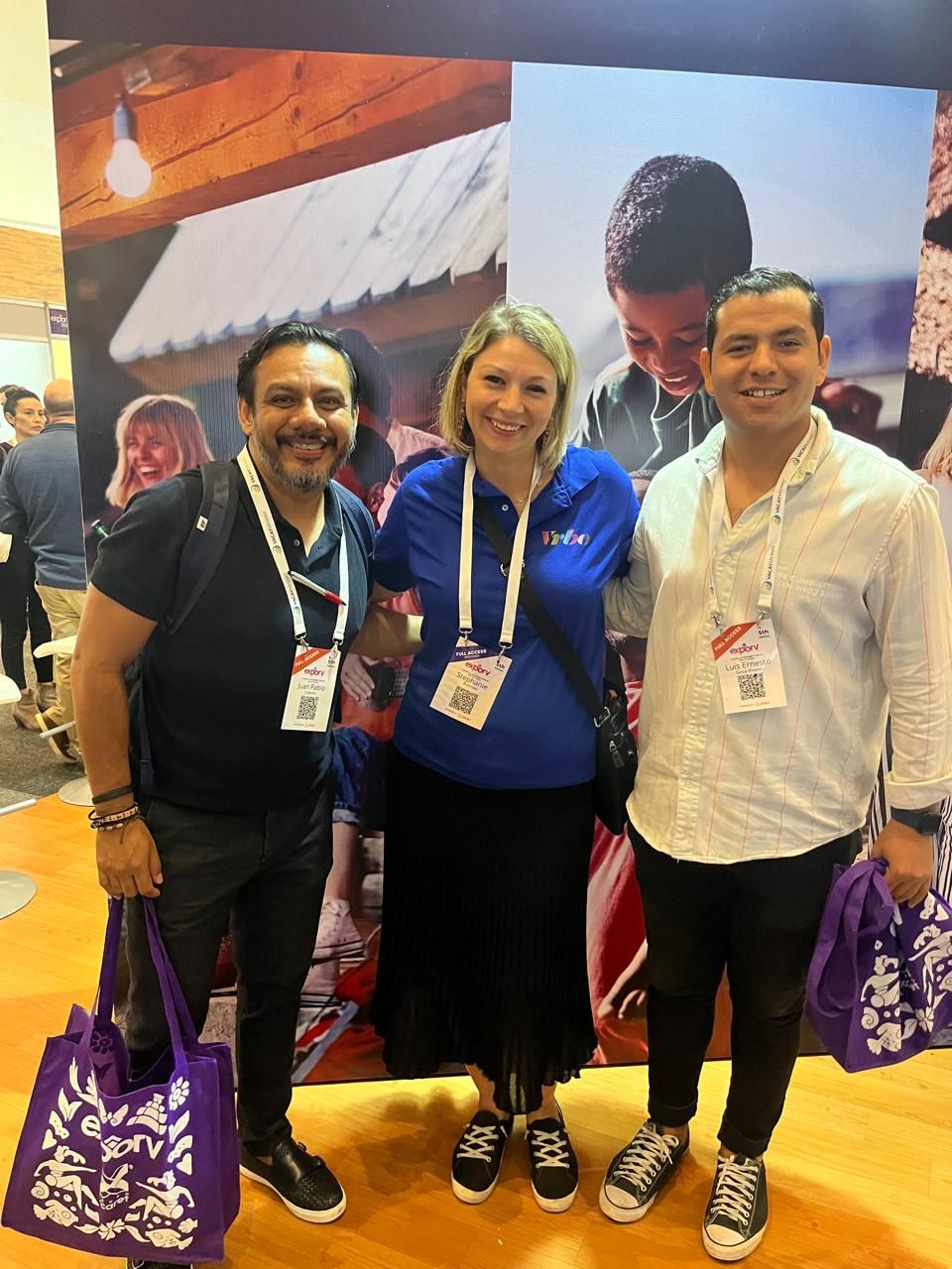 Tres personas posando en un stand de un evento, sosteniendo bolsas de tela moradas y llevando cordones para credenciales.