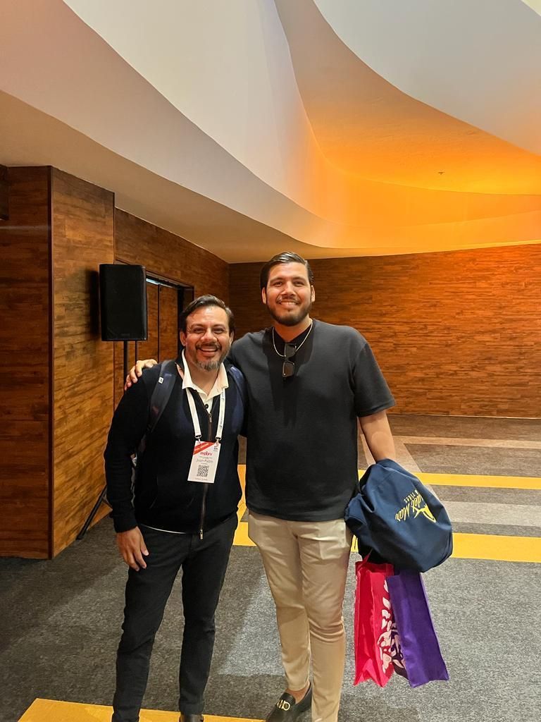 Dos personas de pie en un vestíbulo con paneles de madera, sonriendo, una de ellas sosteniendo bolsas coloridas.