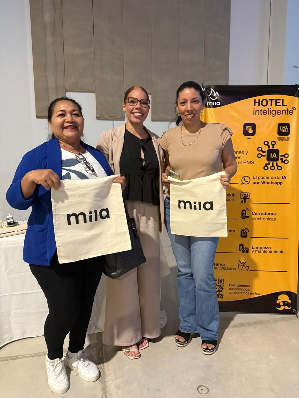 Tres personas posando en el interior con bolsos de la marca Mia junto a un panel amarillo del hotel.