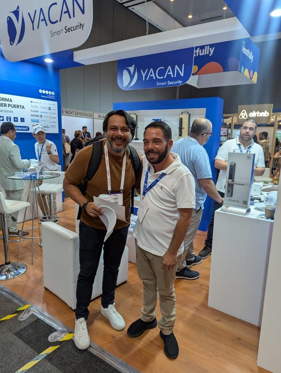 Dos hombres posando juntos en un stand de YACAN, sonriendo en medio de una concurrida feria.