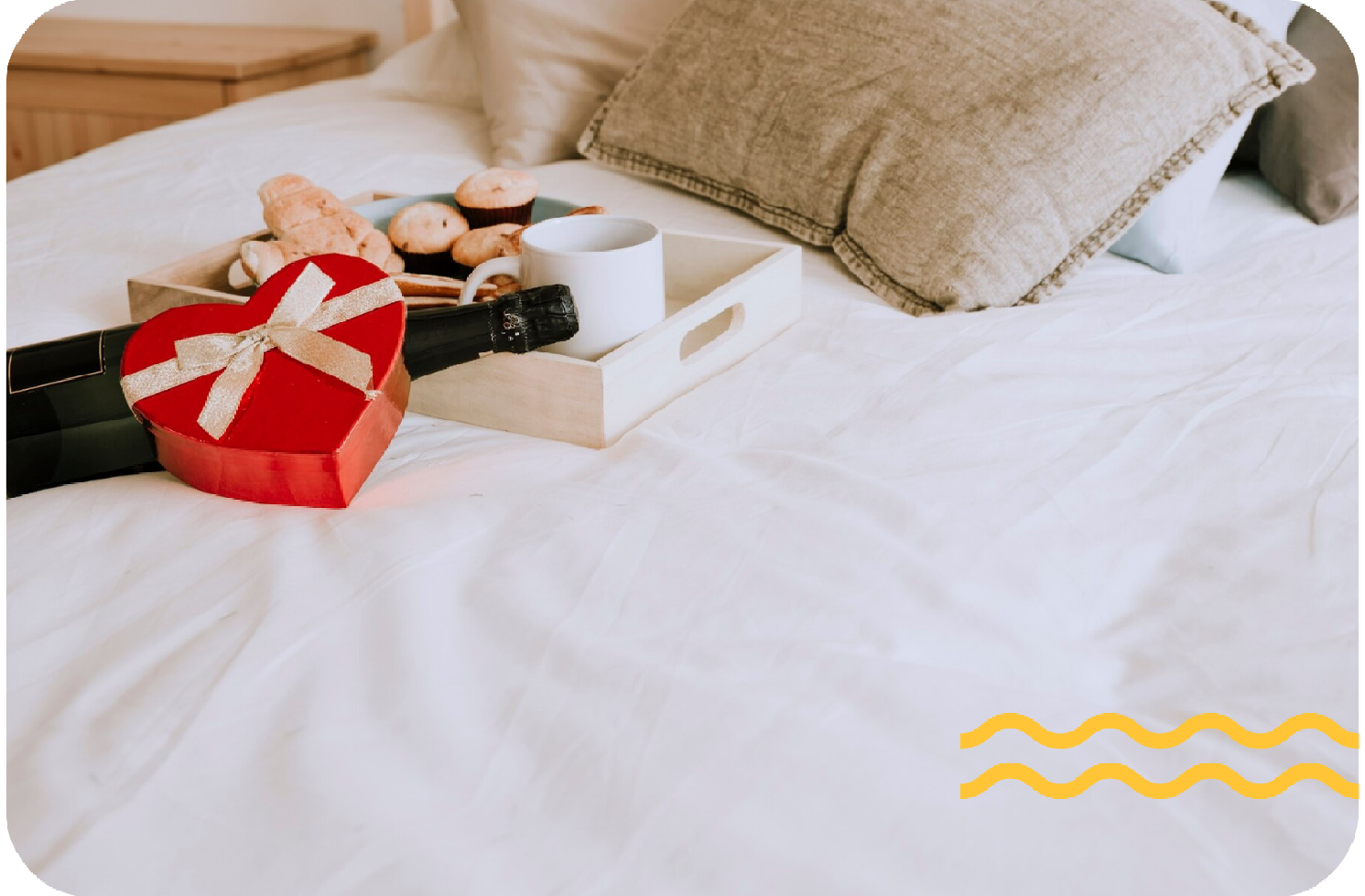 Caja de regalo con cinta y macarons sobre una cama blanca junto a almohadas beige.