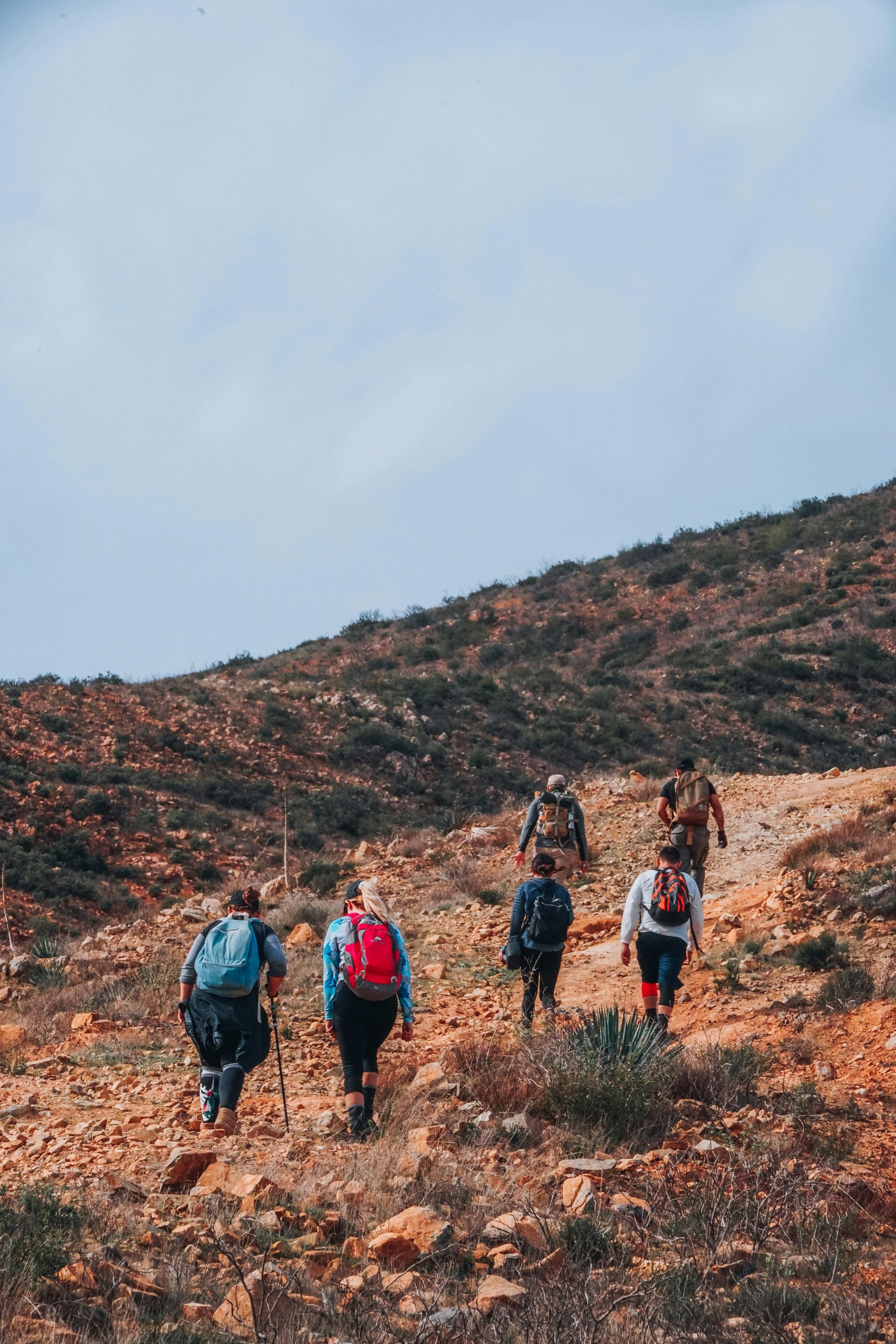 Senderistas con mochilas subiendo una ladera rocosa bajo un cielo nublado.