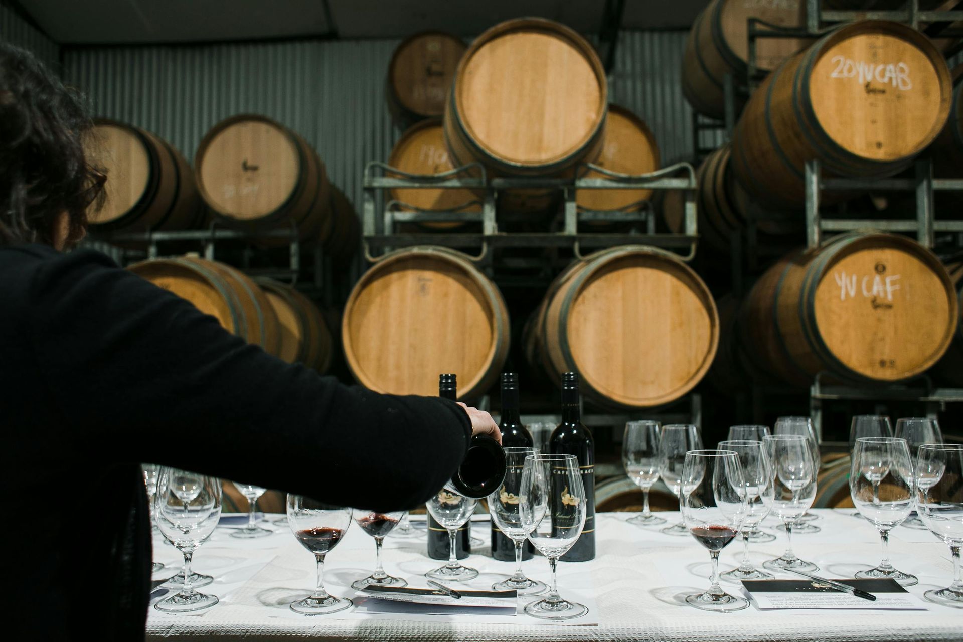 Montaje para cata de vinos con copas sobre una mesa frente a barriles de madera en una bodega.