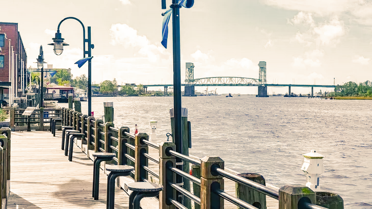 Waterfront scene with a bridge in the distance and a wooden walkway lined with benches.