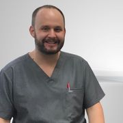 Man with a beard in gray scrubs, smiling, with a pen in his pocket. Neutral background.