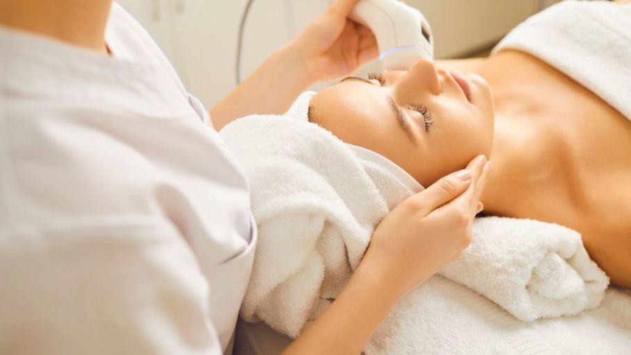 Woman receiving facial treatment at a spa; esthetician using a device on her face.