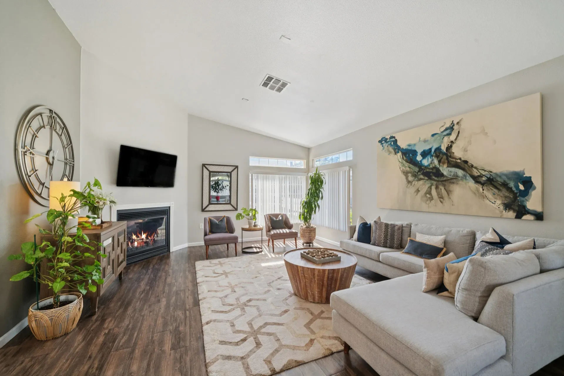 Bright clubhouse room with a gray sectional, fireplace, wall art, and large windows.