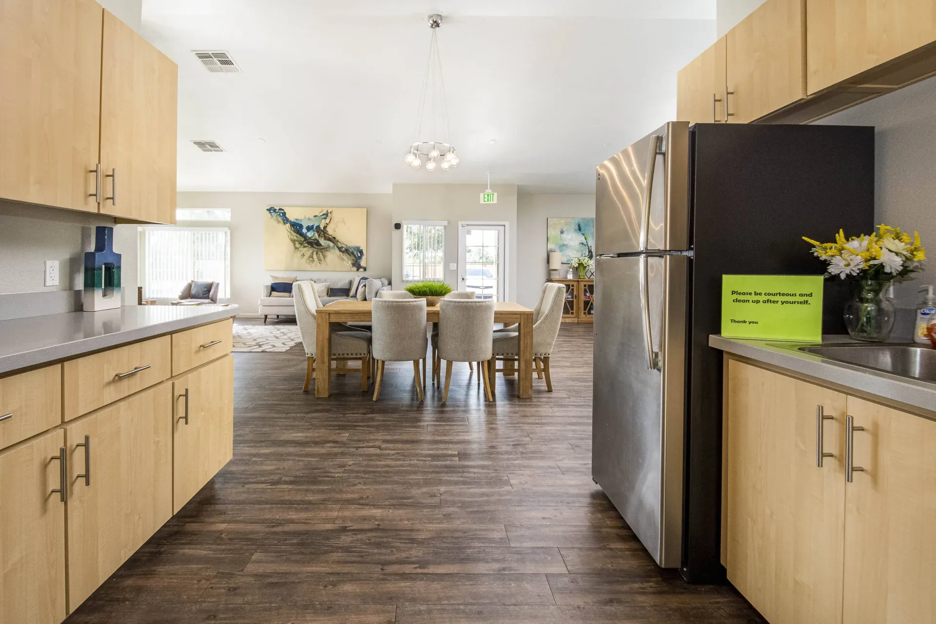 Open-concept clubhouse kitchen with light wood cabinets, gray countertops, stainless steel fridge, and a dining table nearby.