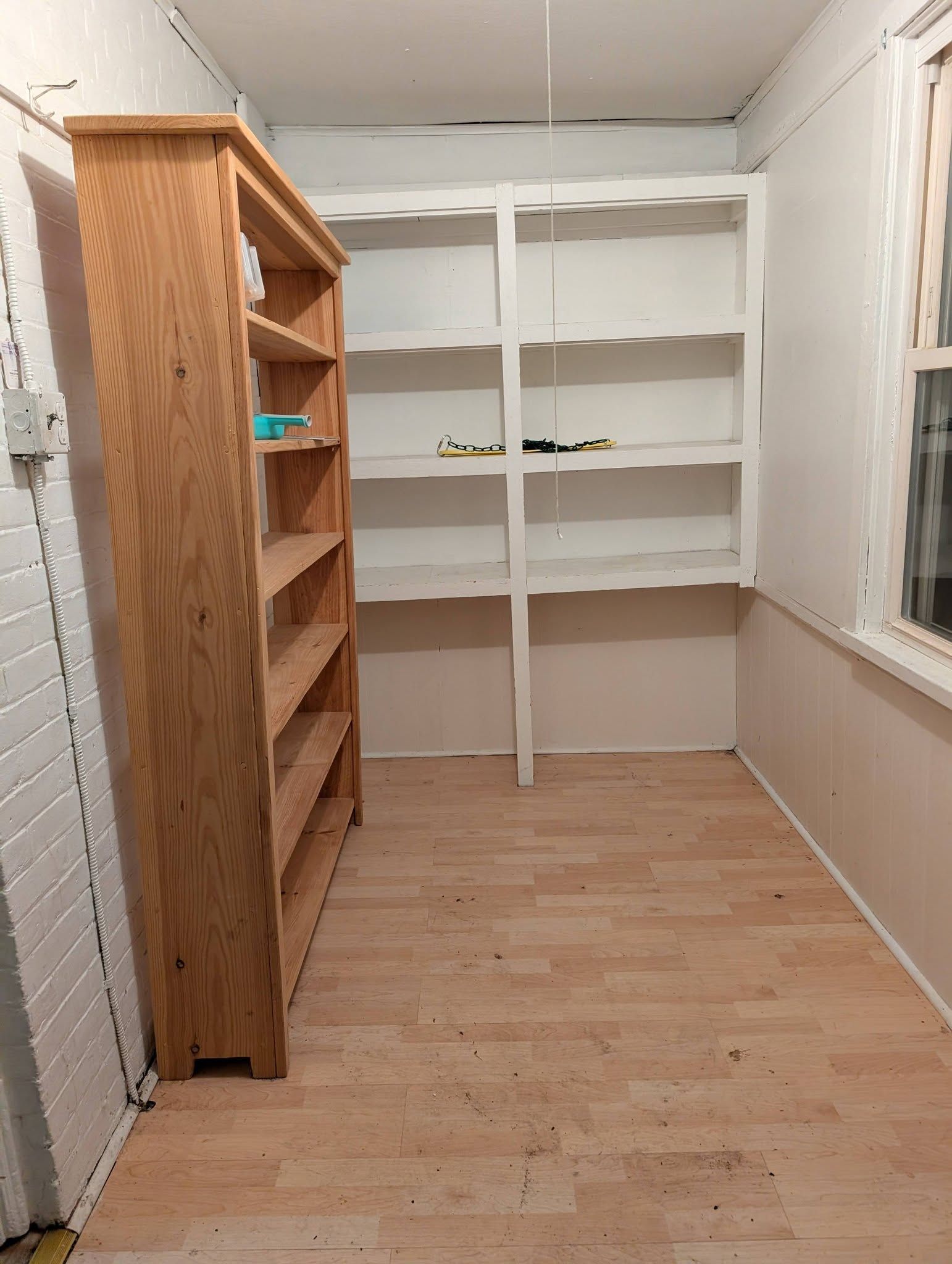 A small pantry or storage room with a wooden bookshelf on the left and white open shelving against the back wall.