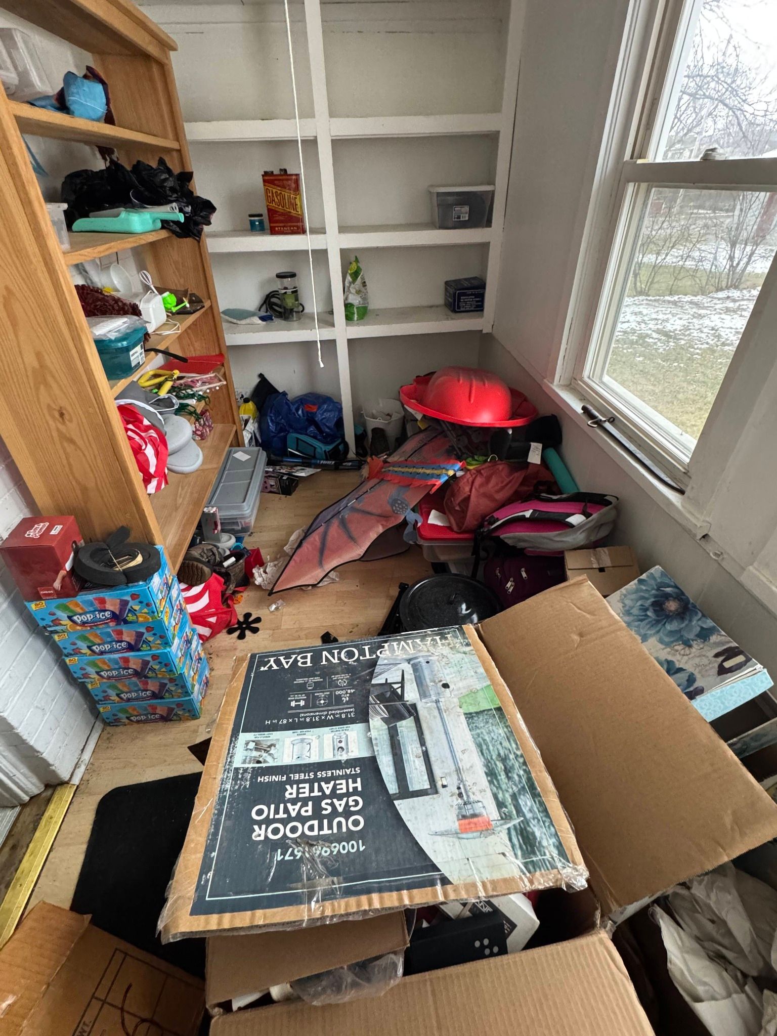 A cluttered storage room with open wooden shelving holding various items, a red play tent, and an open cardboard box.