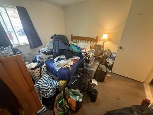 A messy bedroom with laundry, bags, and items piled on and around a bed, illuminated by a lamp on a nightstand.