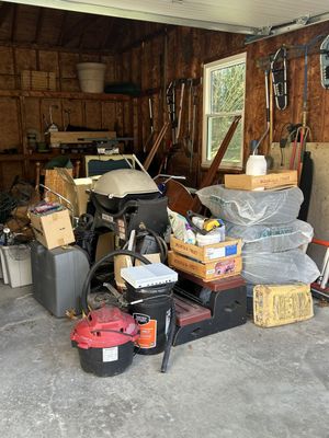 A cluttered garage interior with cardboard boxes, a black shop vacuum, a grill, and items wrapped in plastic.