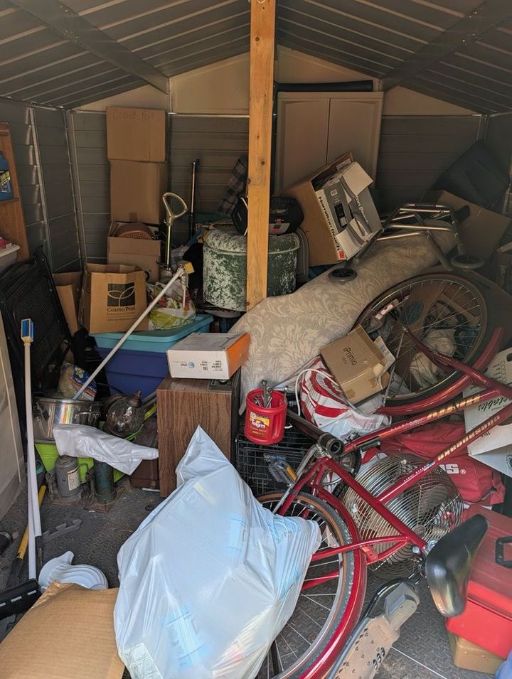 An interior view of a cluttered shed filled with cardboard boxes, a red bicycle, and various household items.