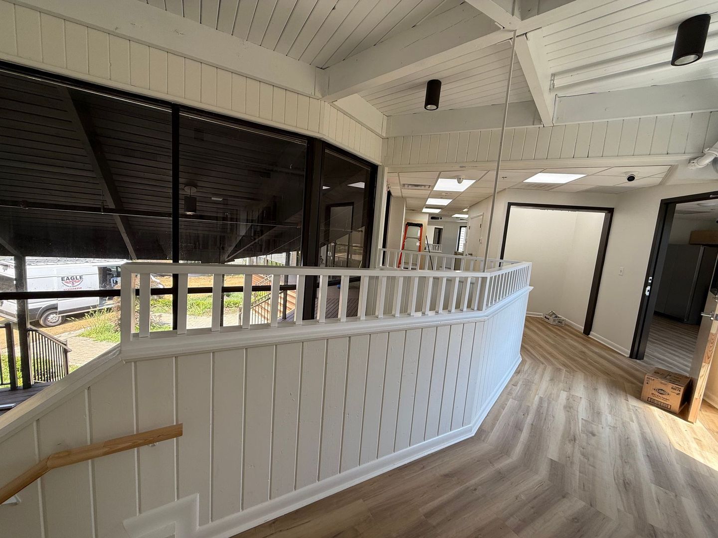 Bright indoor balcony with white railing, wood floor, and black window shades overlooking outside.